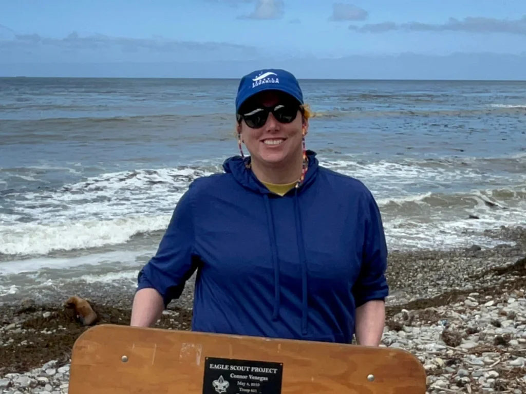 Lindy standing on a beach. She is wearing a blue hat and jacket, and she is holding a wooden herding board.