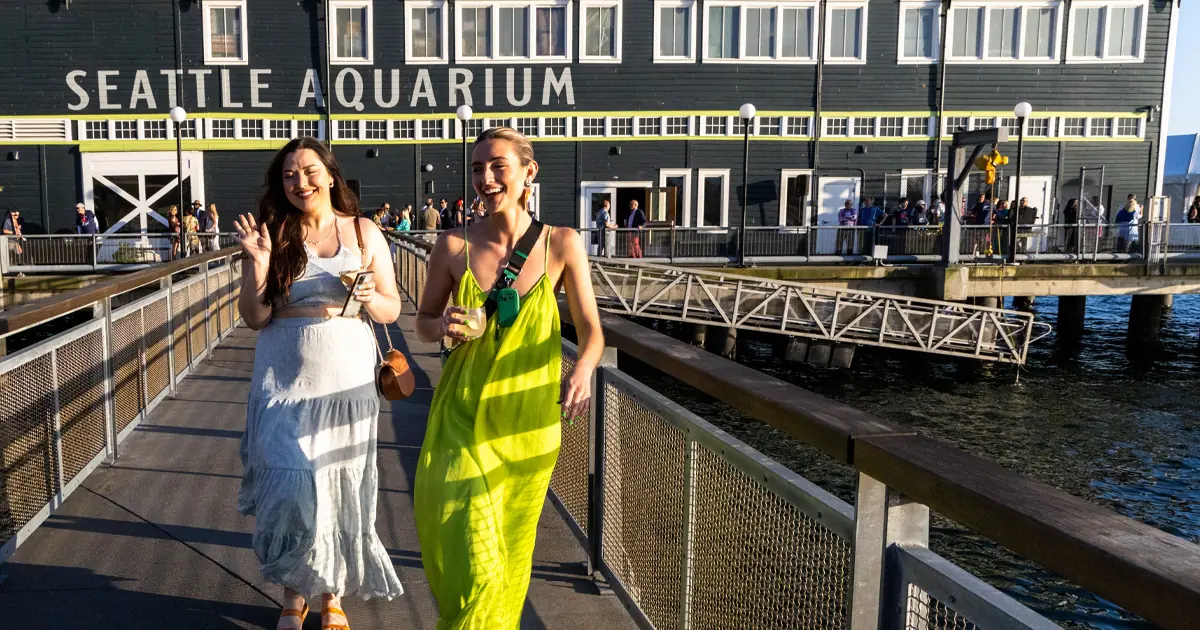 Two adult women, both dressed in casual resort attire, walking along the back pier of the Seattle Aquarium. Both are holding drinks and smiling. The Seattle Aquarium Pier 59 building is behind them.