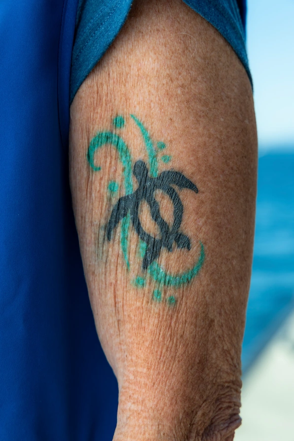 A blue and green tattoo of a turtle done in the style of a Honu petroglyph, located on James' upper left arm.