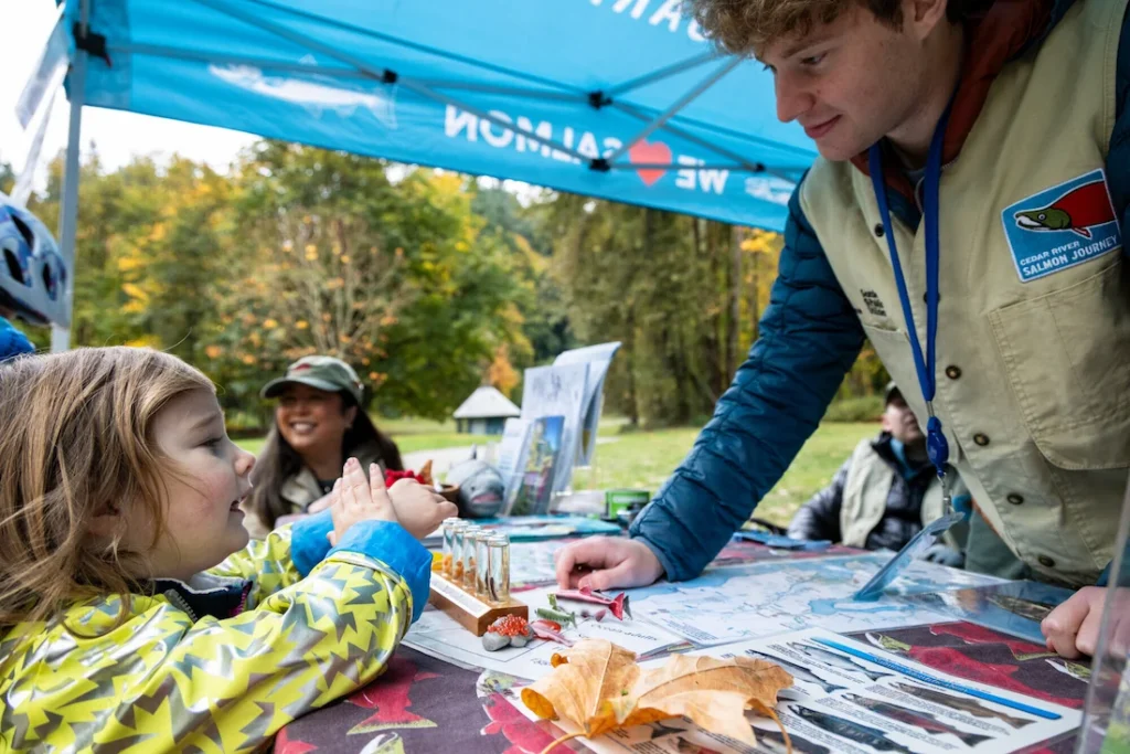 A Seattle Aquarium Cedar River Salmon Journey volunteer interacting with a young girl at a table, with activities and information available across the table to learn about salmon.